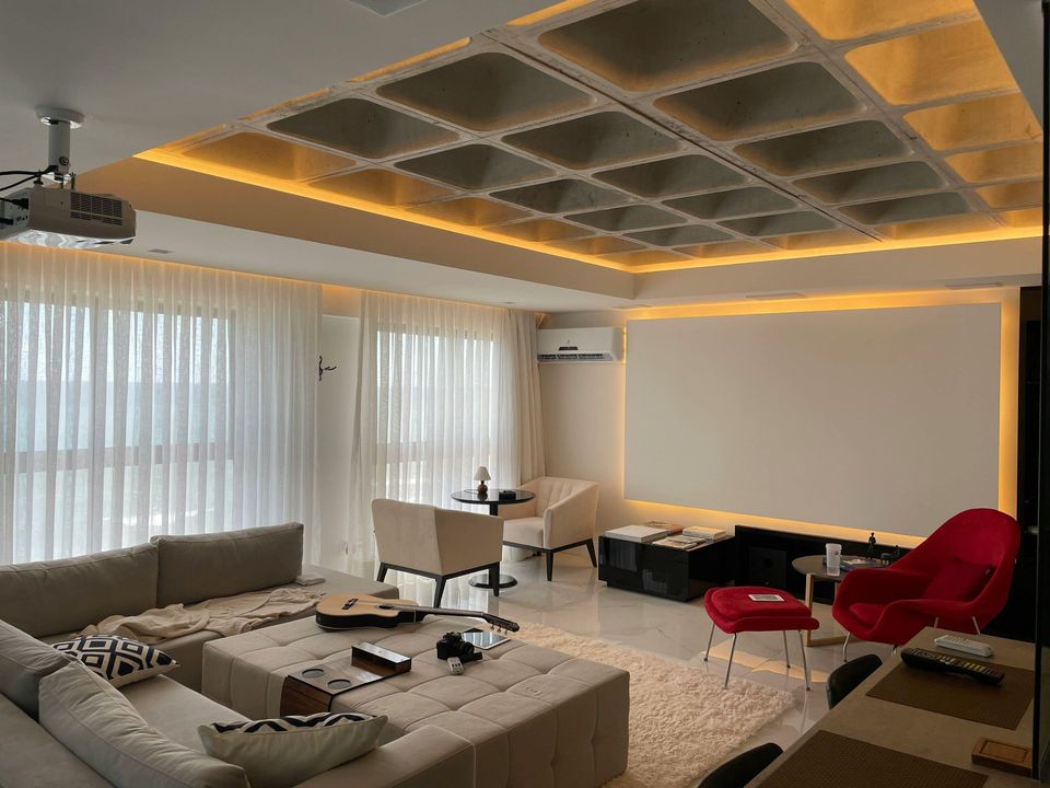 Sala de estar moderna con sofá seccional beige, sillas rojas, grandes ventanales y una pantalla de proyección en blanco.