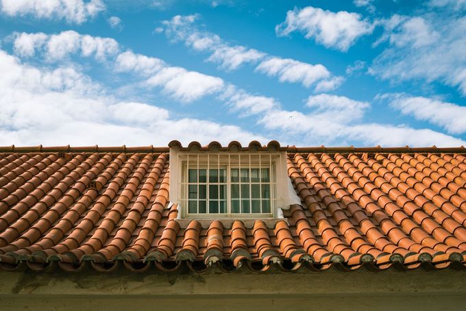 Tejado de tejas de arcilla roja con una pequeña ventana abuhardillada bajo un cielo azul con nubes.