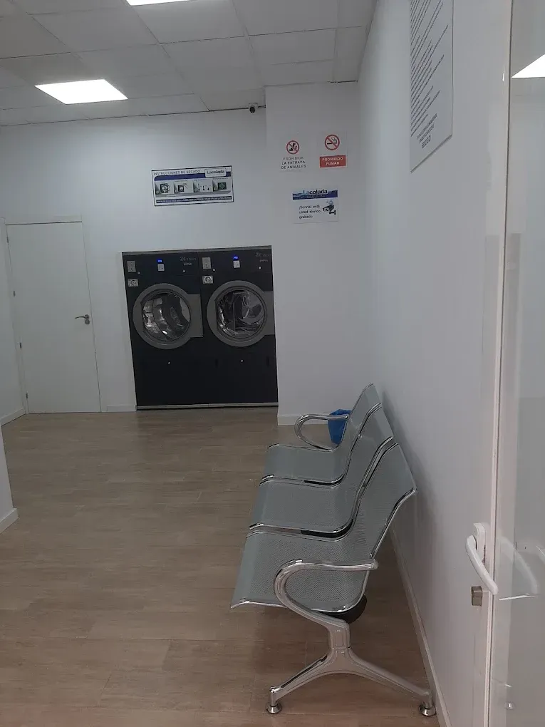 Sala de espera vacía de una lavandería, con sillas grises y lavadoras al fondo