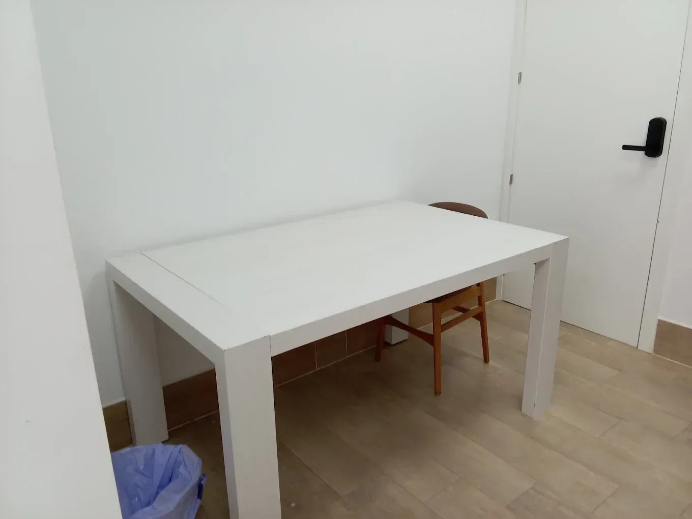 Mesa blanca en una habitación despejada con una silla, una puerta y un cubo de basura azul.