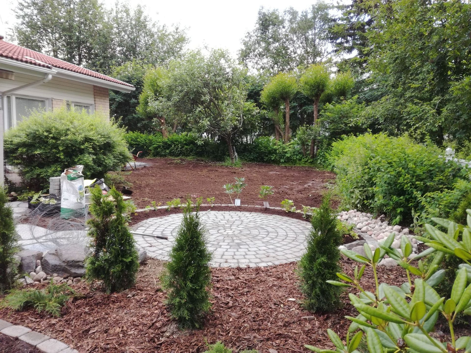 Maisemoitu puutarha, jossa on pyöreä kivinen patio, pensaita ja vihreitä puita talon vieressä