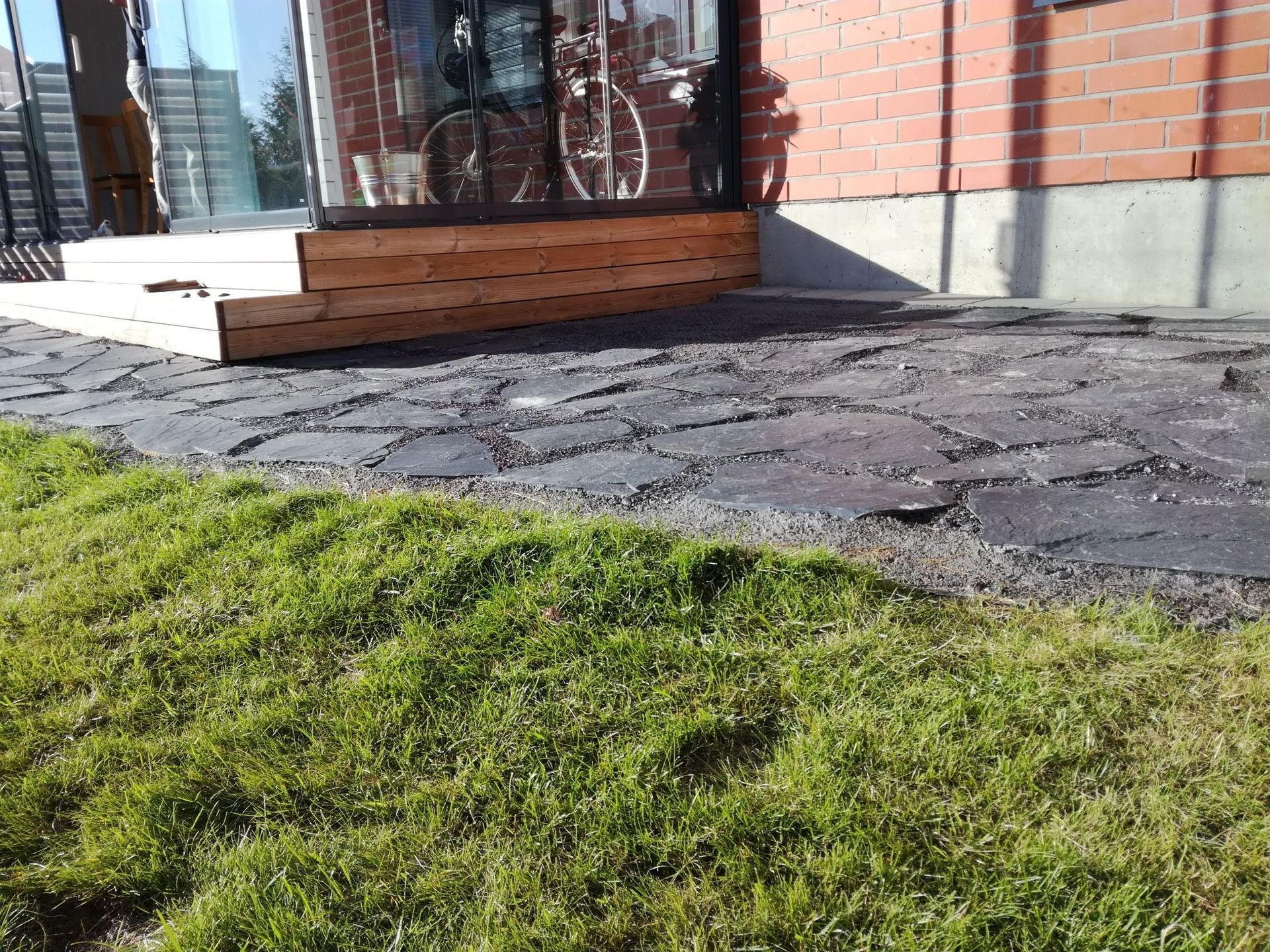 Vihreä nurmikko halkeilevan jalkakäytävän ja rakennuksen sisäänkäynnin vieressä, jossa on puiset portaat ja lasiovet