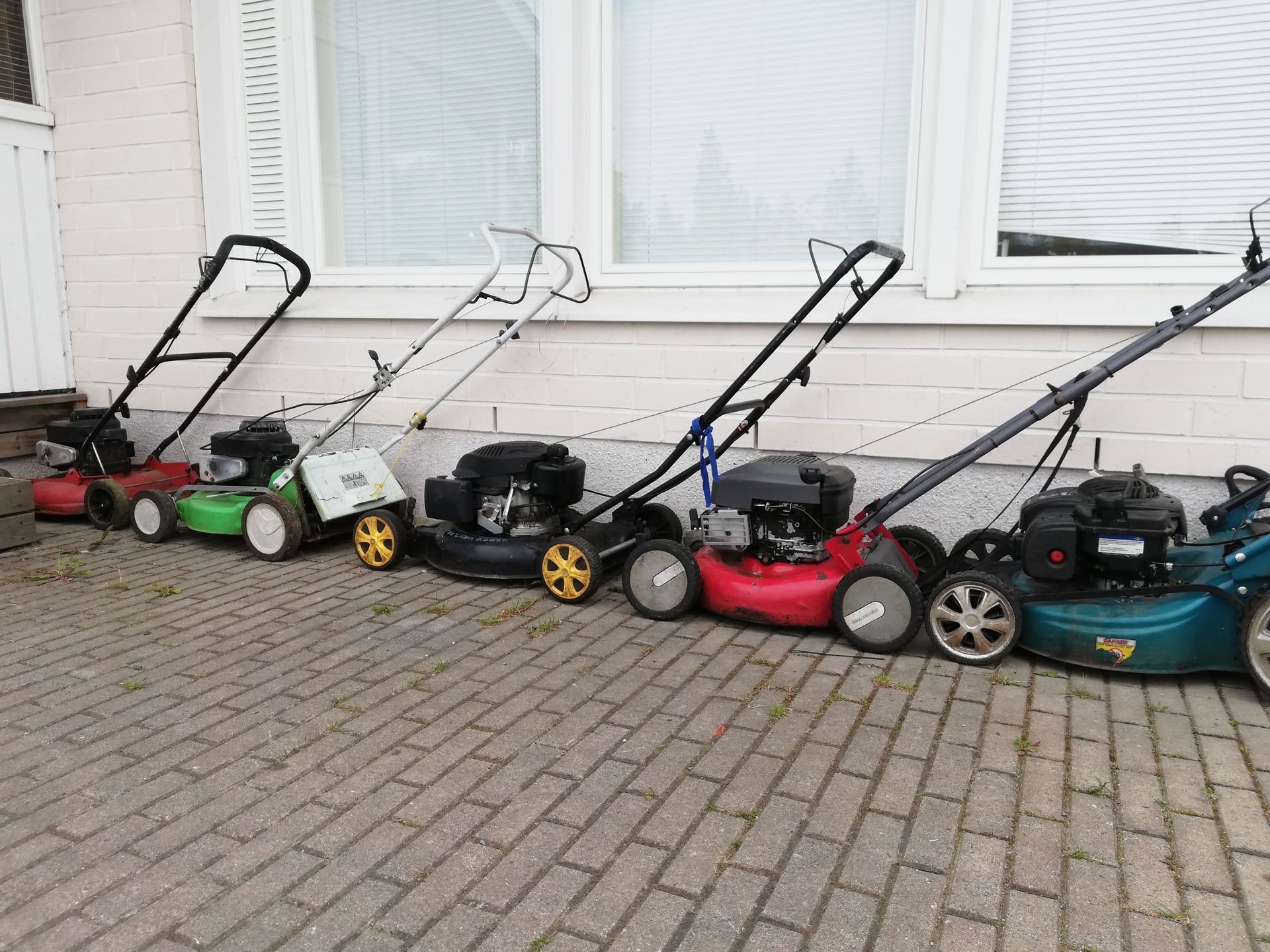 Viisi ruohonleikkuria rivissä tiilillä päällystettyllä ajotiellä valkoisen talon edessä.
