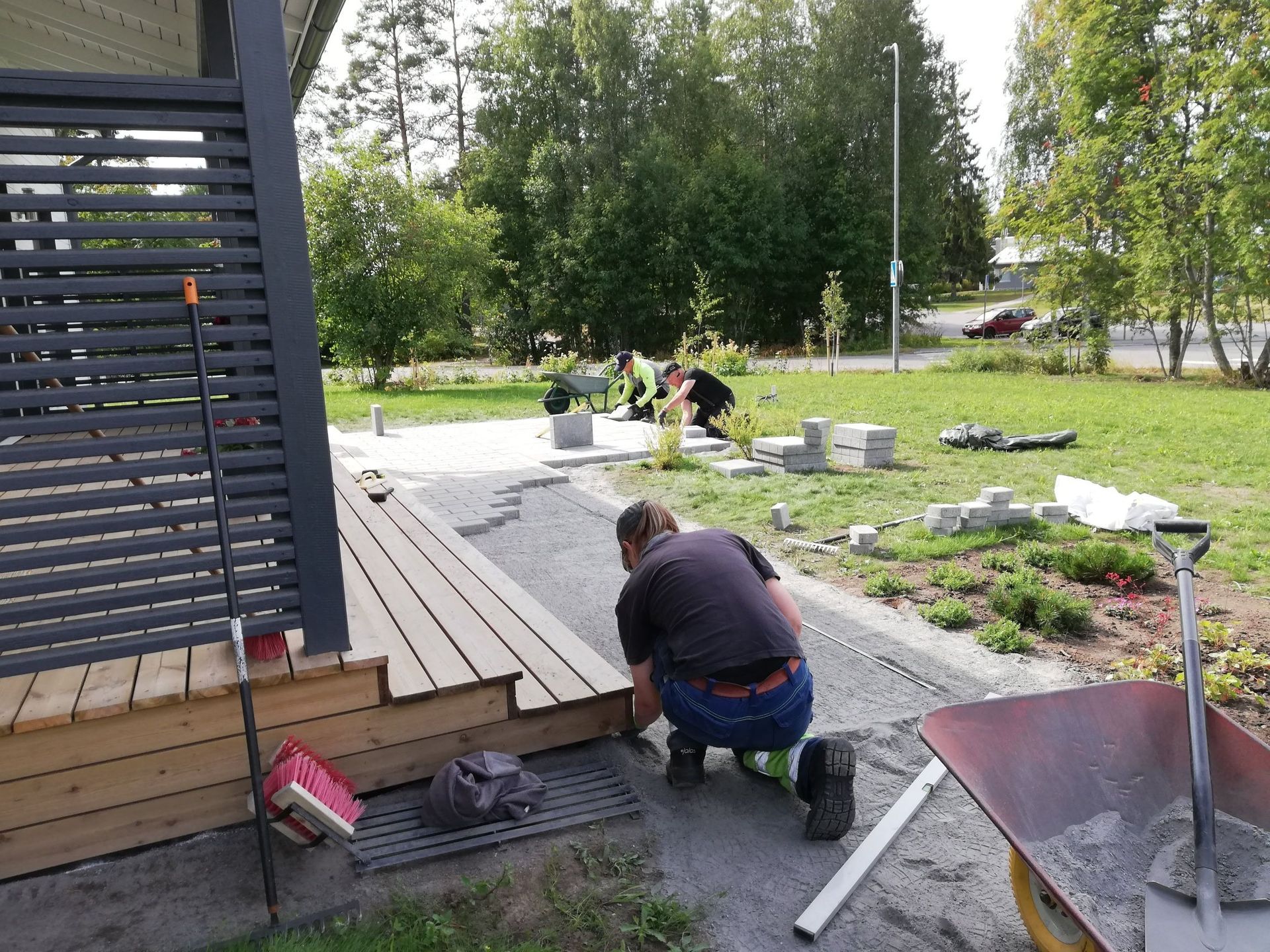 Työntekijä polvistuu terassikannella puutarhapolun vieressä ja asentaa lautoja työkalujen ja materiaalien lähelle
