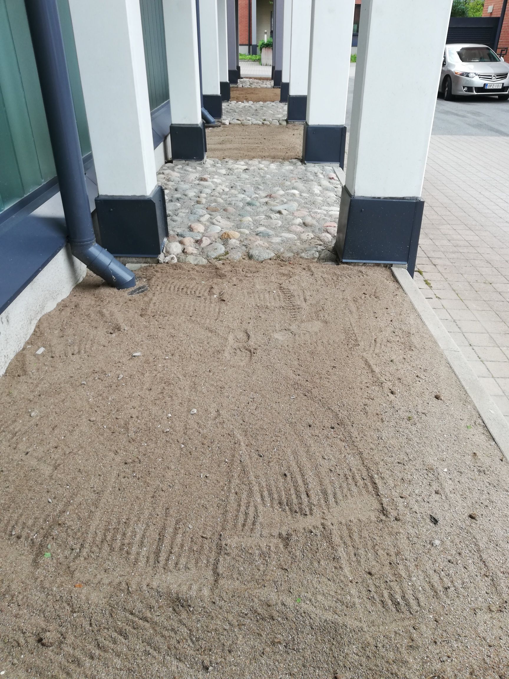 Valkoisten pylväiden välissä kulkeva sorakäytävä johtaa päällystetylle kadulle, taustalla pysäköity auto.