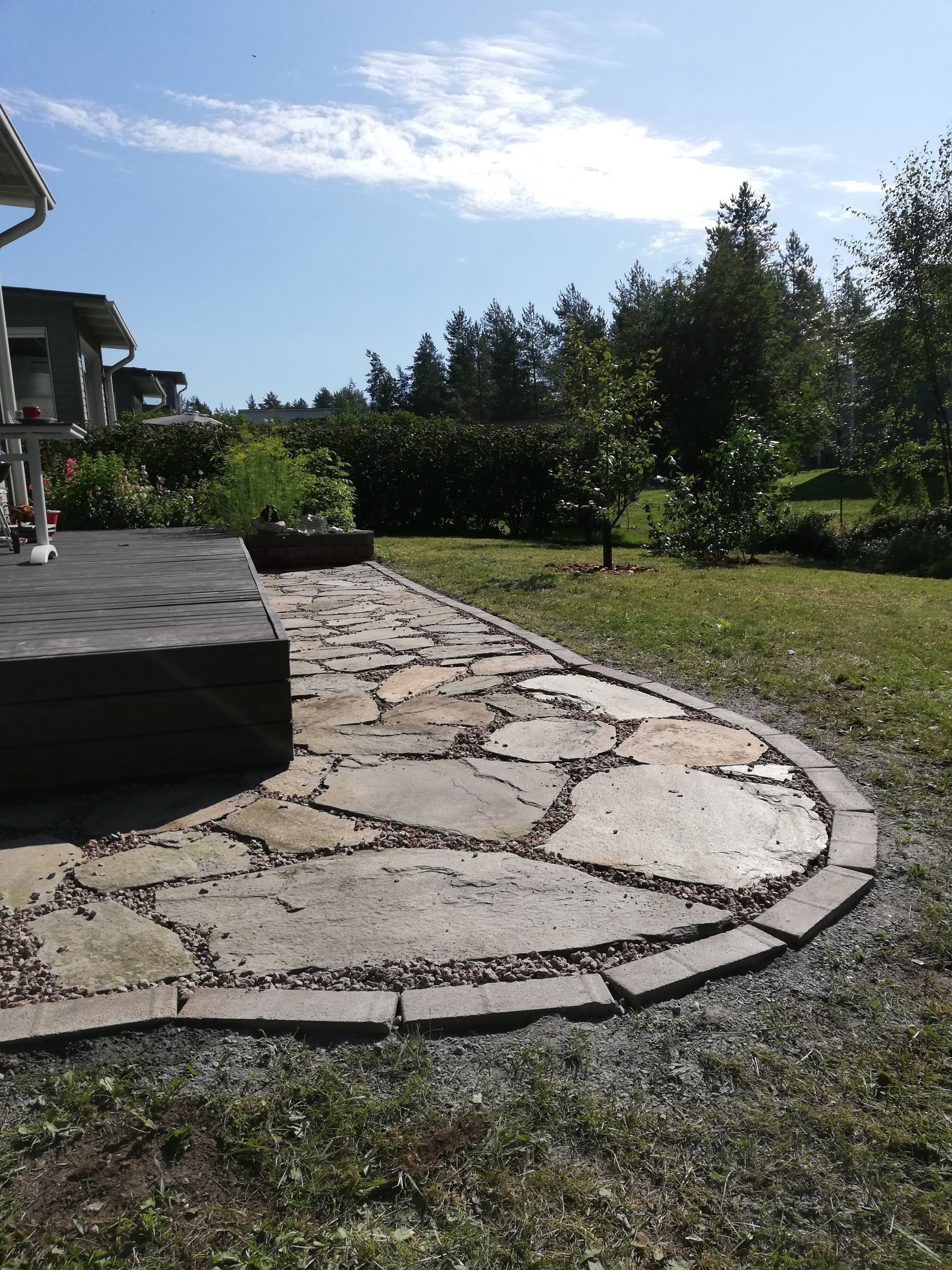 Kivinen patio, joka kaartuu aurinkoisen takapihan läpi, jossa on nurmikkoa, puita ja vasemmalla talon kuisti