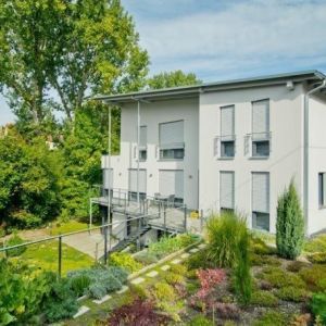 Helle Fassade mit grauen Fensterrahmen und Jalousien vor Bäumen.