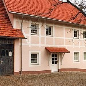 Teilansicht eines Hauses mit hellrosa Fassade, weißen Sprossenfenstern und einem tiefgezogenen roten Ziegeldach.