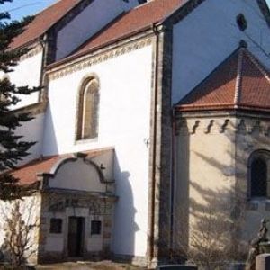 Nahaufnahme einer weißen Kirchenwand mit dekorativem Gesims und angrenzendem niedrigerem Dachbau.