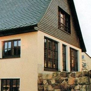 Hauswand mit grünen Schindeln im Giebelbereich und Sprossenfenstern.