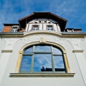 Untersicht einer hellen Fassade mit floralem Stuckdekor, einem Rundbogenfenster und dunklem Zierfachwerk im Giebelbereich.