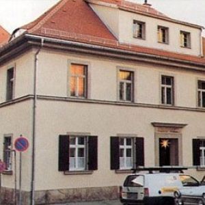 Eckansicht eines mehrstöckigen, hellen Wohnhauses mit klassischer Fensteranordnung unter einem rötlichen Ziegeldach.