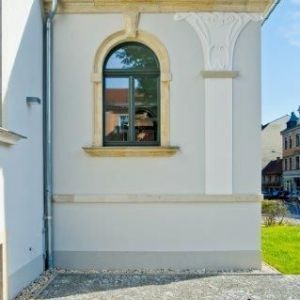 Nahaufnahme eines tiefgrünen Rundbogenfensters in einer hellen Fassade mit verzierten Kapitellen und Stuckelementen.