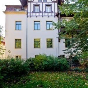 Frontalansicht eines hellgelben Gebäudes mit regelmäßiger Fensteranordnung und einem hölzernen Zwerchgiebel im Landhausstil.