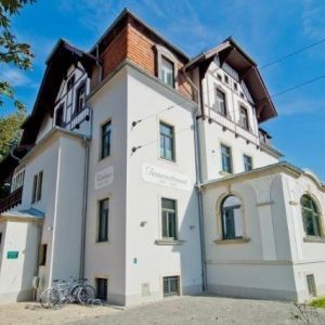 Außenansicht einer weißen, mehrstöckigen Villa mit markantem dunklen Fachwerk im Dachgeschoss und einem hölzernen Balkon.