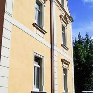 Seitenansicht einer gelben Fassade mit Fokus auf die Fenstergiebel und das herabführende Regenrohr vor blauem Himmel.