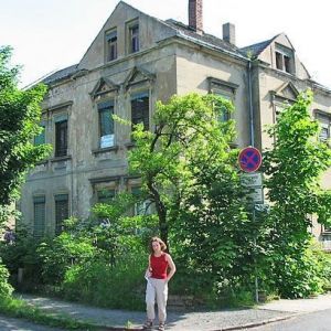 Fassade eines mehrgeschossigen, unsanierten Gebäudes in einer Innenstadt, teilweise durch grüne Bäume verdeckt.