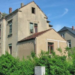 Ein sanierungsbedürftiges, graues Gebäude mit abblätterndem Putz und teilweise eingeschlagenen Fenstern vor blauem Himmel.
