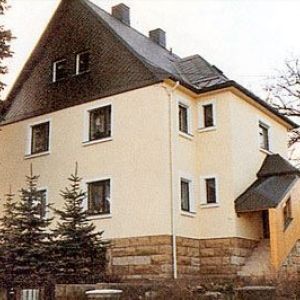 Ein Wohnhaus mit hellgelber Fassade und einem markanten, steilen Krüppelwalmdach, das mit dunklen Schindeln verkleidet ist.