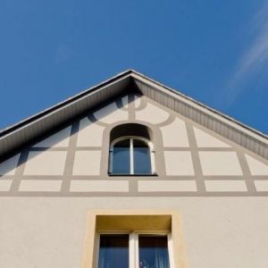 Detailansicht eines Giebels mit dekorativem grauen Fachwerk-Muster auf weißem Grund und einem zentralen Rundbogenfenster.