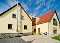 Außenansicht eines modernen, hellgelben Einfamilienhauses mit Satteldach und roten Dachziegeln unter blauem Himmel.