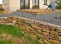 Rustikale Trockenmauer aus Natursteinen zur Hangbefestigung in einem Garten mit Blick auf eine Holzterrasse.