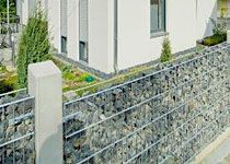 Moderner Zaun aus Gabionen (Steinkörbe) und Betonpfeilern als Grundstücksbegrenzung vor einem Wohnhaus.