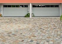 Zwei weiße Sektionaltore einer Doppelgarage vor einer gepflasterten Hofeinfahrt aus Naturstein.