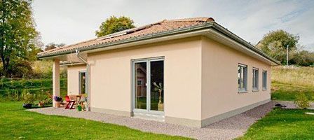 Hell verputzter Winkelbungalow mit großen Glasfronten und Kiesumrandung im Garten.