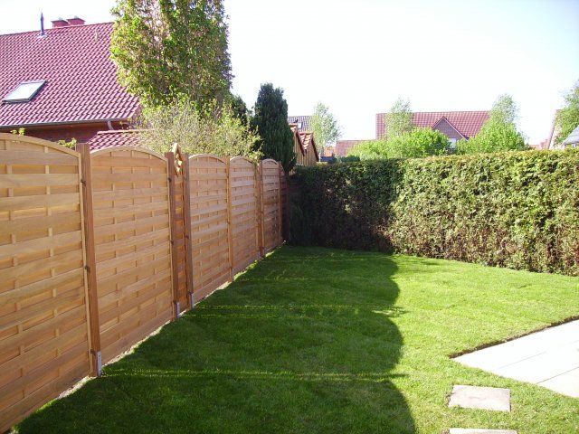 Ein Hinterhof mit Holzzaun und grünem Rasen, im Hintergrund eine Hecke und ein Haus. Sonniger Tag.