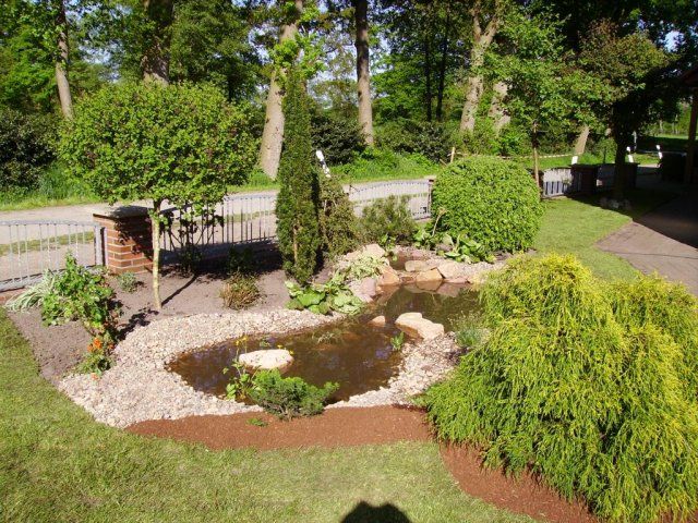 Landschaftlich gestalteter Garten mit Teich, Sträuchern, Bäumen und Kiesrand. Umgeben von grünem Rasen.