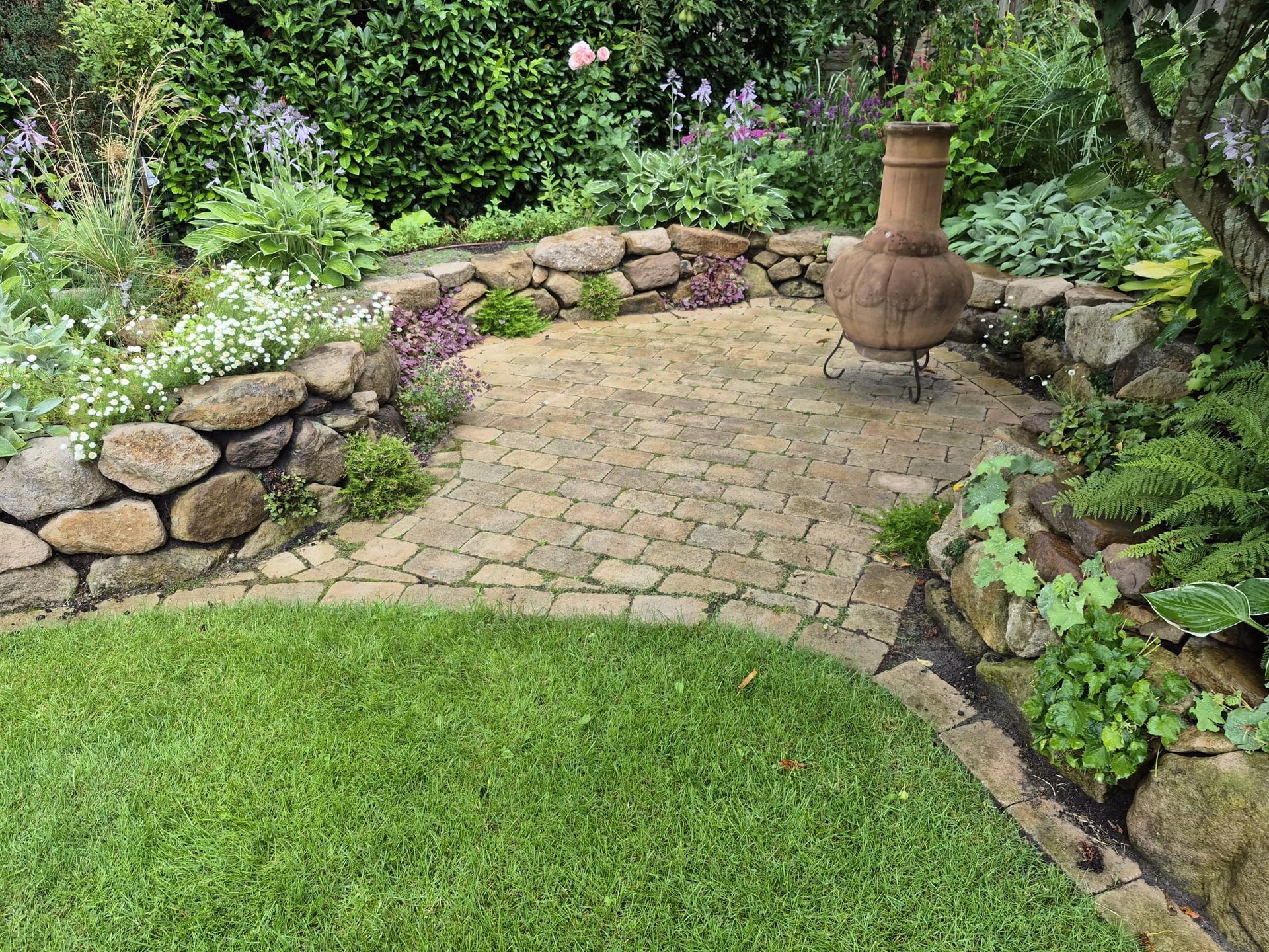 Eine mit Ziegeln gepflasterte Terrasse mit Steinmauern, Rasenfläche und Topfpflanzen in einer Gartenanlage.