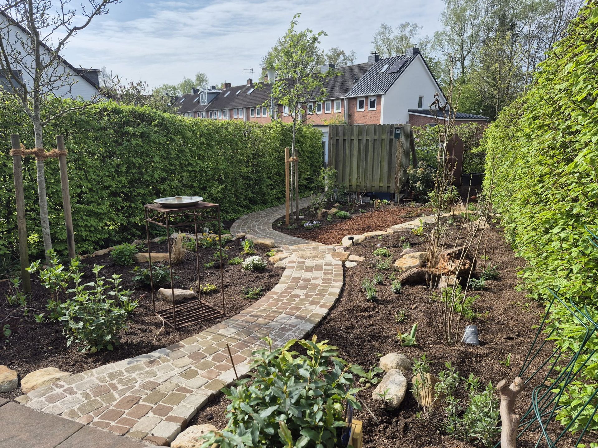 Ein Backsteinweg schlängelt sich durch einen Garten mit Hecken, Bäumen und dunklem Boden. Im Hintergrund sind Häuser zu sehen.