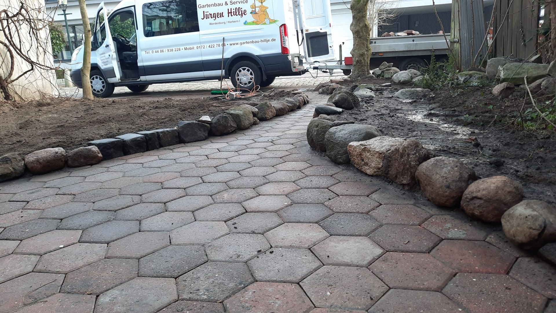 Sechseckige Pflastersteine bilden einen von Steinen eingefassten Gartenweg. Ein weißer Lieferwagen parkt in der Nähe.