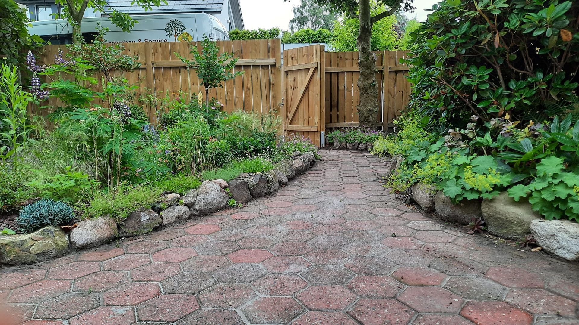Ein Ziegelweg führt zu einem Holztor in einem Garten, der von Pflanzen und einer Steinumrandung gesäumt ist.
