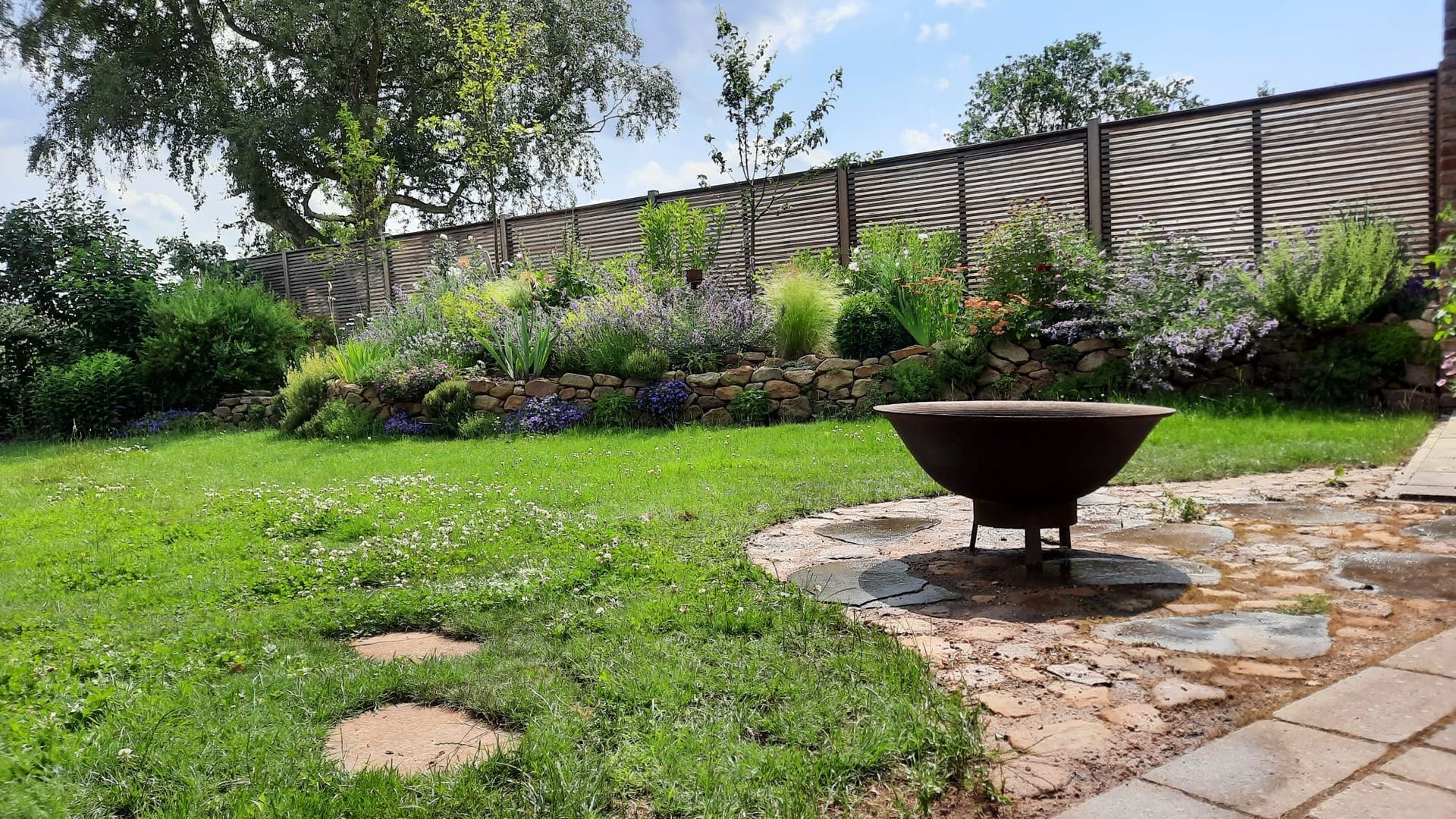 Üppig grüner Hinterhof mit Steinterrasse, Feuerstelle und einem Hochbeet an einem Holzzaun.