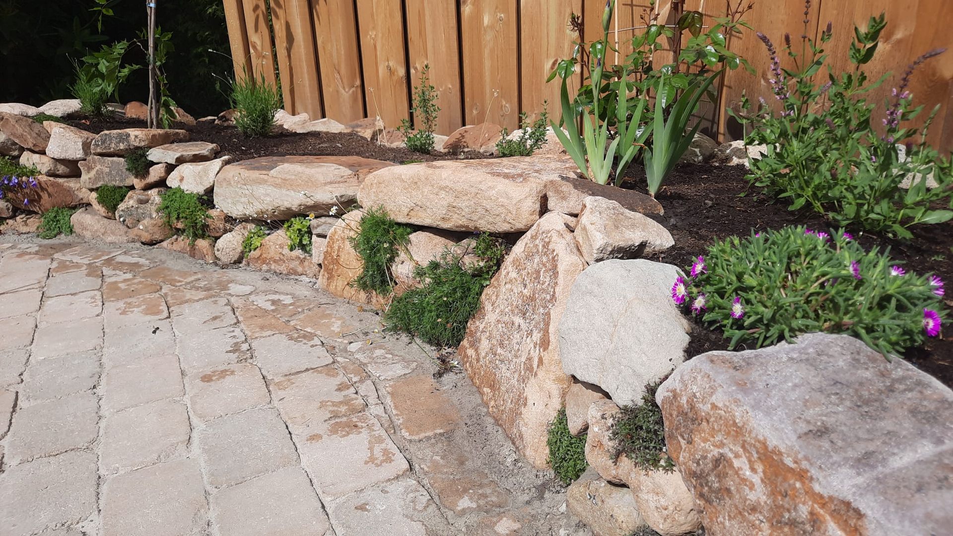 Steinmauer mit gepflastertem Gehweg davor, Garten mit Pflanzen und Holzzaun im Hintergrund.
