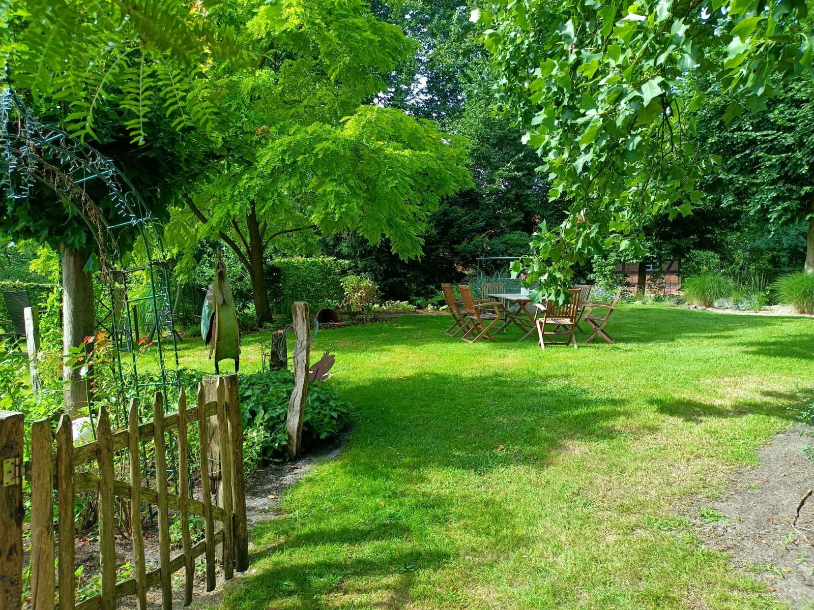 Grüner, sonnendurchfluteter Hinterhof mit Holzzaun, Rasen, Bäumen und einem Tisch mit Stühlen.
