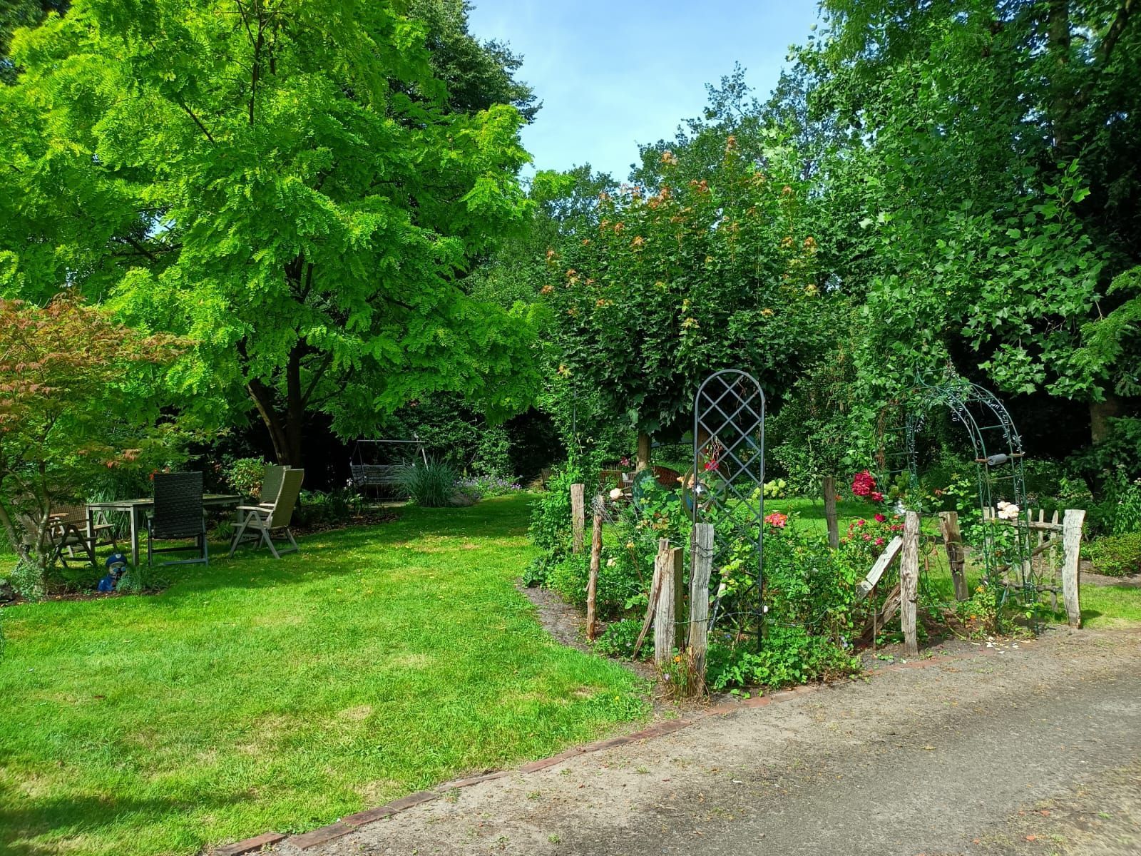 Ein üppiger Garten mit grünem Gras, Bäumen und einem Holzzaun, der einen Weg säumt. Unter den Bäumen stehen eine Bank und Stühle.