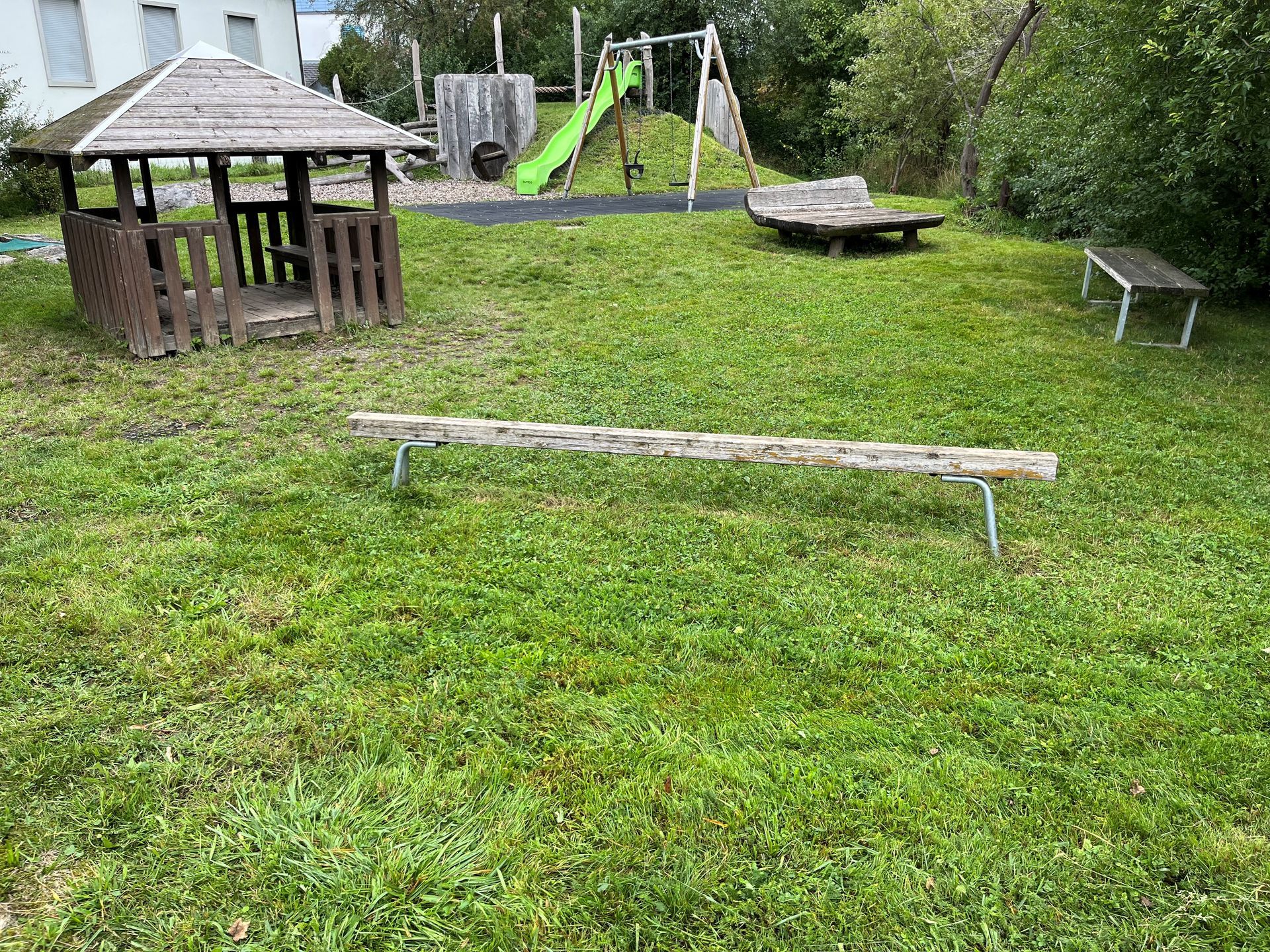 Eine Holzbank steht inmitten einer üppigen grünen Wiese neben einem Spielplatz.