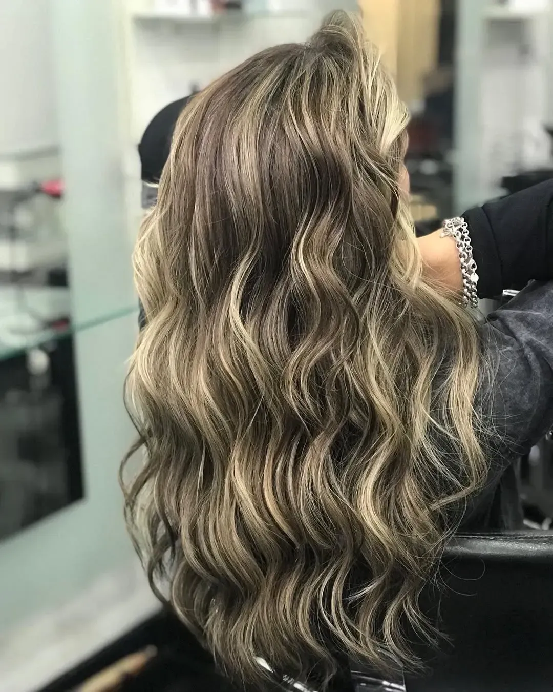 Cabello largo, ondulado, castaño oscuro con reflejos rubios más claros, peinado en rizos sueltos en un salón de belleza.