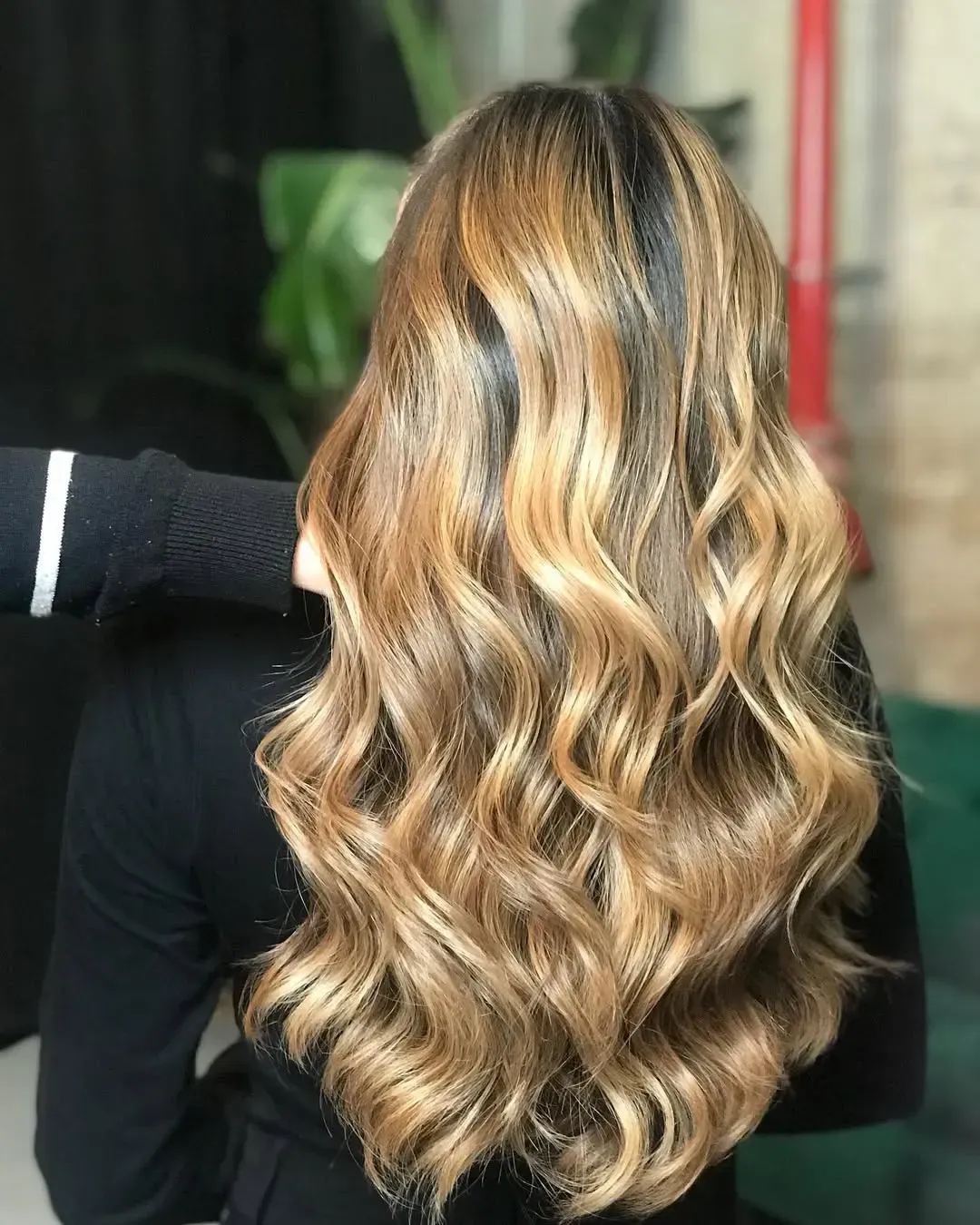 Cabello largo y ondulado con reflejos en tonos miel y caramelo, peinado en rizos sueltos sobre un fondo oscuro de interior.