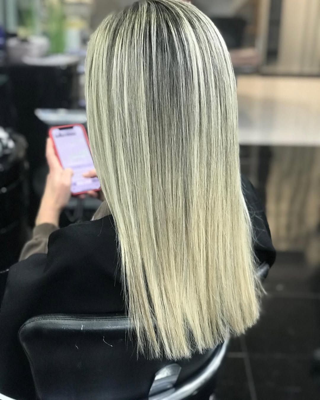 Una persona sentada en una silla de peluquería, de espaldas a la cámara, con el pelo largo, liso y rubio con mechas.