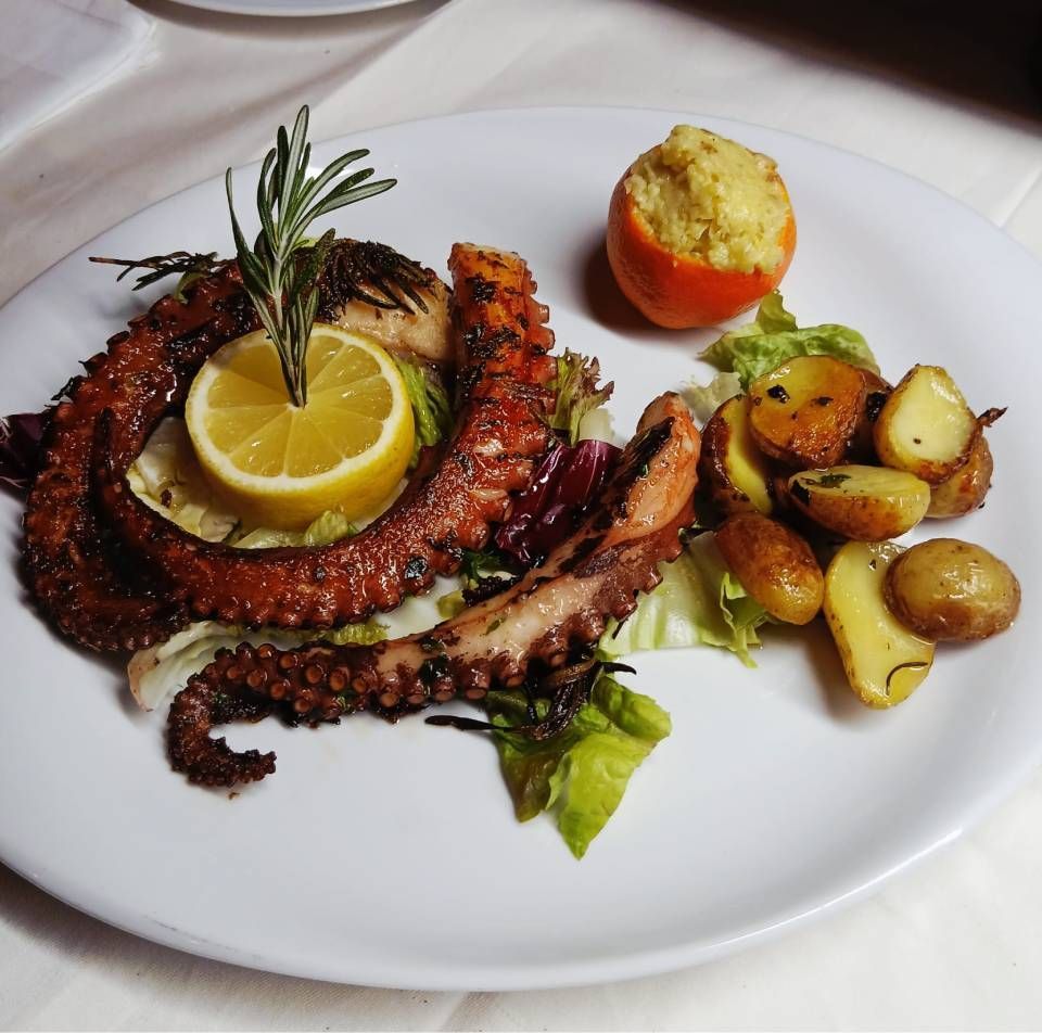 Gegrillter Oktopus mit Zitrone, Rosmarin, Kartoffeln und einer gefüllten Tomate auf einem weissen Teller.