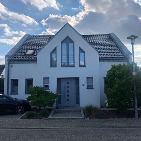 Weißes zweistöckiges Haus mit grauem Dach, grauer Haustür und grauen Fenstern; kleine Bäume davor, Auto links.