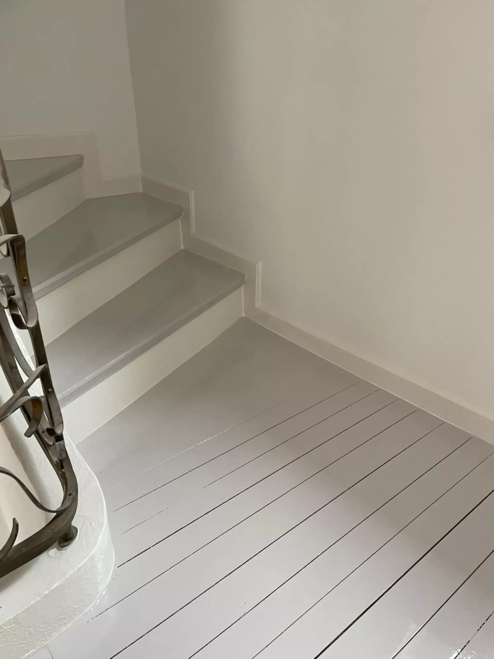 Treppenhaus mit hellgrauer Treppe und hellgrauem Boden. Weiße Wände. Links ein schmiedeeisernes Geländer.