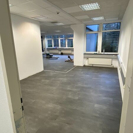 Leerer Raum mit dunkelgrauem Boden und weißen Wänden, Fenstern und Deckenleuchten.