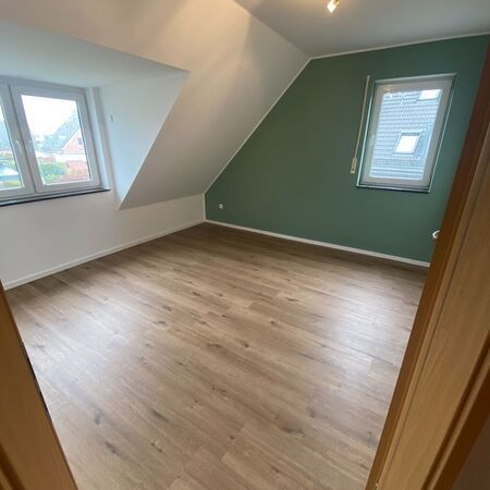 Leerer Raum mit schrägen Wänden, Holzboden, zwei Fenstern und einer grünen Wand.