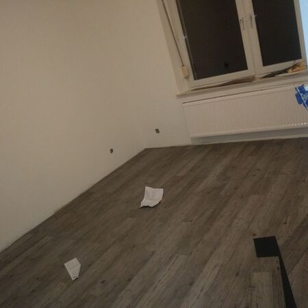 Leeres Zimmer mit neu verlegtem Holzboden, weißen Wänden und einem Fenster mit einem Heizkörper darunter.