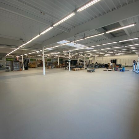 Große, leere Lagerhalle mit hellem Deckenlicht und grauem Boden. Kisten und Möbel stehen überall verstreut.